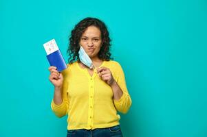 Upset and sad young woman traveler in yellow sweater puts her medical mask off and grimaces looking at camera, holding a boarding pass, air flight ticket and passport. Copy ad space. Travel concept photo