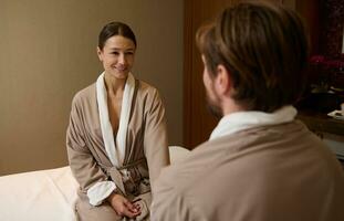 Happy delighted beautiful young woman in bathrobe smiles looking at her husband, holding hand, relaxing at spa resort on honeymoon. Cute couple getting beauty treatment together in the wellness center photo