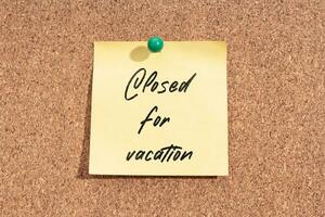 Yellow sticky note with message Closed for Vacation on cork board photo