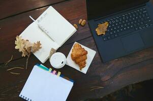 Flat lay autumnal composition with open notepad and copybook, laptop with fallen oak leaves, takeaway paper cup with hot drink and delicious baked croissant on a wooden table in the forest cafe photo
