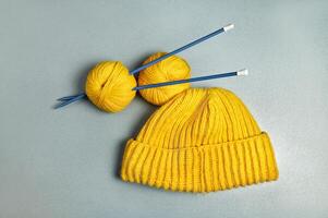 An item photograph of a yellow knitted woollen hat and two winding wool threads strung on long blue knitting needles on a grey background photo