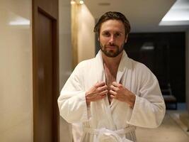 Attractive middle aged man with a charming look and a slight unshaven, handsome macho wearing white terry bathrobe, standing in a lobby of a luxury wellness spa complex, looking at camera photo