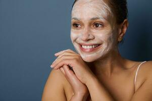 Attractive woman smiles with beautiful toothy smile, posing against gray background with facial mask, removing make-up and cleaning her face using exfoliant facial cleansing beauty cosmetic product photo