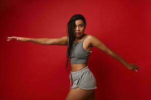 African American sportswoman, determined athlete with stylish dreadlocks stretches her hands, taking them to the sides, looks at the camera, stretching out on a red background with copy space photo