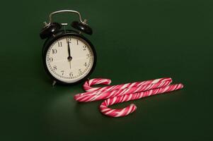 Studio shot with soft shadows of a black alarm clock with midnight on the clock face and sugary striped Christmas lollipops, candy canes isolated over dark green background with copy space for ad. photo