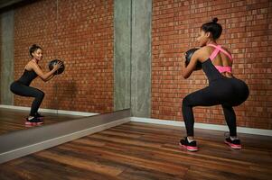 Rear view of attractive slim fitness African woman doing glute exercises, squatting with medical gymnastic ball looking at her mirror image while working out photo