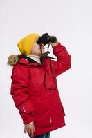 Adorable curious elementary aged Caucasian boy in yellow hat and bright red down jacket, looking through binoculars, isolated on white background, copy ad space. Adventure and Winter travel concept photo