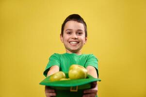 A happy boy holds a leprechaun hat full of apples out in front of him to the camera. Isolated on yellow background. Saint Patrick day photo