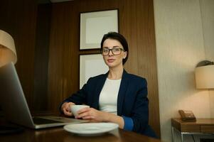 Confident portrait of a prosperous businessman, successful female sales manager looking at camera while sitting at the table and drinking coffee in her office and writing a progress report on laptop photo