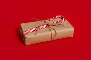 Beautiful minimalist composition of Christmas items, gift box wrapped in kraft wrapping gift paper with tied bow and sweet candy cane isolated over red background with copy space for advertisement photo