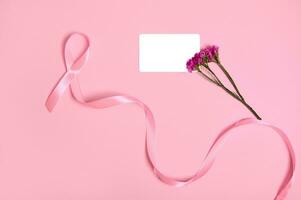 Flat lay of a long pink ribbon with endless one end, symbol of Breast Cancer Awareness Day, a cute bouquet of flowers and an empty white blank plastic card with copy space, on pink background. photo