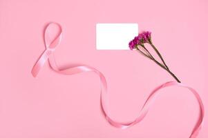 Flat lay of a long pink ribbon with endless one end, symbol of Breast Cancer Awareness Day, a cute bouquet of flowers and an empty white blank plastic card with copy space, on pink background. photo