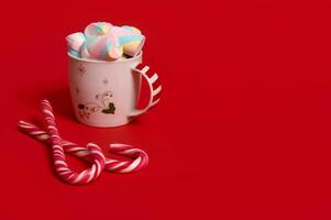 Studio shot of a mug of hot drink with marshmellows and candy canes on red colored background with copy space for Christmas advertising photo