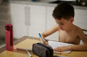 Adorable child boy draws anime comics on white paper sheet. Pencil case and water bottle on marble table photo