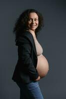 auténtico grávido mujer, participación su embarazada barriga, sonriente mirando aparte, vestido en elegante chaqueta de sport, aislado en gris foto