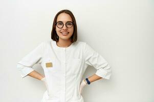 sonriente joven médico interno en blanco médico Saco posando con brazos en cintura en contra un blanco antecedentes con Copiar espacio foto