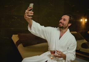 Handsome attractive man in bathrobe sitting by the pool in wellness spa centre, holding cup of tea and smiles happily making self-portrait or talking by video call while getting relax in spa resort photo