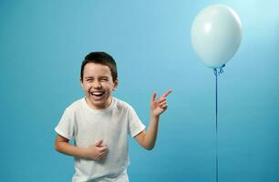 Cute boy laughing and pointing finger up on a balloon , looking at camera standing against a blue background photo