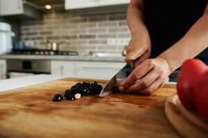 Close-up of hands of a woman chopping black olives on a wooden board photo