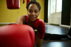 africano americano hermosa atlético mujer, hembra Boxer en rojo boxeo guantes estiramientos el piernas en el enroscarse en el piso de un Deportes gimnasio y puñetazos hacia cámara. marcial combate Arte y extensión foto