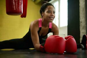 Close-up of African female athlete, boxer in boxing red gloves performing a twine on the floor of a sport gym with a boxing punching bag. Martial combat art stretching, sport and wellness concept photo