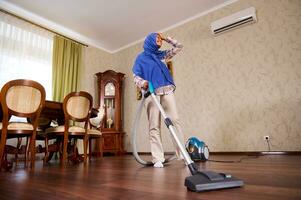 Delightful happy Arab Muslim young woman or housewife with vacuum cleaner, vacuuming the floor at home, dancing, singing, enjoying cleanup. Household, housework and cleaning concept. photo