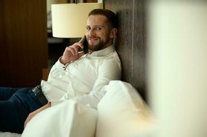Charming young European man, businessman, talking on mobile phone, smiling with a beautiful toothy smile, looking at camera, resting after business meetings and negotiationson a bed in a hotel bedroom photo