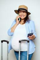 Smiling Latin American pregnant woman holds a passport document, talks on mobile phone, booking hotel, isolated on white photo