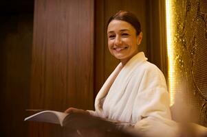 Close-up of middle aged elegant Caucasian woman, attractive brunette dressed in white terry bathrobe, smiles toothy smile looking at camera, reading magazine while resting at wellness spa lounge area photo