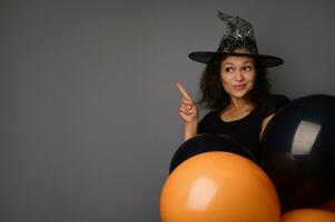 Mysterious mixed race woman dressed in witch carnival costume holds colorful orange and black air balloons, points on a copy space on gray background. Concept of Halloween traditional seasonal event photo