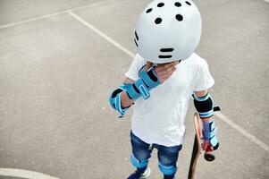 Adolescente chico en protector casco y engranajes participación un patineta, mirando abajo y limpiando lágrimas desde su ojos foto