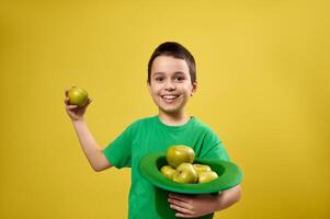 Funny little boy stands isolated on yellow background and holds an Irish leprechaun hat full of apples in one hand and a green apple in the other. Saint Patrick's day photo