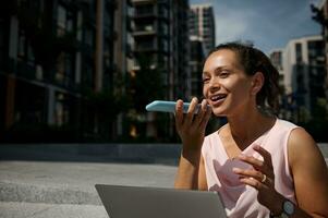 Adorable mixed race beautiful woman recording a voice message on smartphone while working on laptop sitting on steps on the buildings background photo