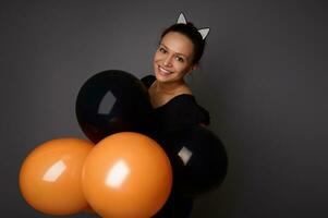 African American woman dressed in black, wearing hoop with cat ears, smiles toothy smile holds black and orange air balls in front of her, looks at camera against gray background. Halloween concept photo