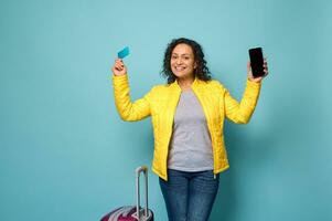 Cheerful woman in bright yellow jacket, with luggage raises her arms up, holding mobile phone with blank space and plastic credit or discount card, isolated over blue background with copy ad space photo