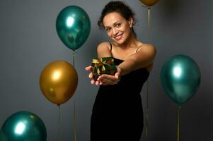 Mixed race cute woman in black dress holds a Christmas gift in outstretched hands and shows it, looking at camera, isolated over gray background with gold and green air balloons, copy space for ad photo