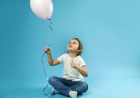 Smiling little girl sitting on blue background with soft shadow and holding balloons in the hand. Childhood and child protection day concept. Copy space photo