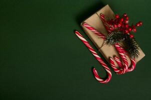 Flat lay of beautiful Christmas present in craft wrapping paper decorated with holly and sweet striped white and red lollipops candy canes laid out in the corner of copy space on dark green background photo