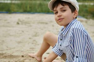 Adorable schoolboy looking at camera while playing on the sand during summer vacations. Summer camp. Headshot portrait. photo