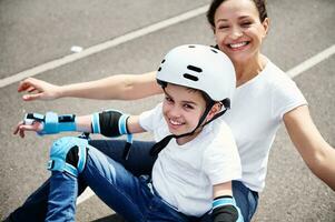 Happy mom and adorable son in protective helmet and equipment enjoying time together sitting on a skateboard photo