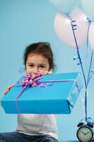 atención en azul regalo con un rosado arco, cuales un linda pequeño niña sostiene fuera a el cámara mientras sentado en un azul antecedentes. celebracion concepto foto
