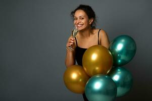 Attractive Hispanic woman smiles looking at camera, poses with glass of sparkling wine and luxury festive shiny golden and green metallic air balloons against gray wall background with copy space photo