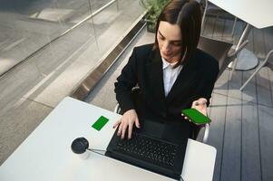 Top view of woman holding smartphone with chroma key green screen, typing on laptop keyboard sitting in a cafe at a table with coffee cup and green blank credit discount card with copy ad space photo