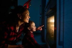 Happy loving mother with her adorable daughter in red and green plaid shirt sitting on the floor and looking through the oven at the traditional German Stollen bread baked on a baking tray photo