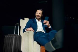 Handsome businessman, investor, entrepreneur sitting in an armchair, resting in a hotel lobby with a smartphone in his hands and crossed legs and smiling, looking to the side during a business trip. photo