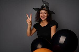 Beautiful smiling with toothy smile woman wearing wizard hat, dressed in witch carnival costume for Halloween party, shows a peace sign, poses against gray background with black balloons, copy space photo