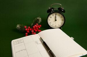 Close-up of an opened page of notepad with plans for day, alarm clock and snowy branch of holly Christmas plant isolated on green background photo