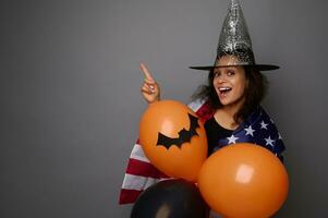 Attractive mixed race woman in wizard hat, wrapped in American flag, holds colorful orange air balls, smiles toothy smile looking at camera, points on copy space on gray background. Halloween concept photo