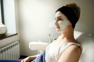 Side portrait of a woman sitting on a chair in spa salon with beauty cleansing mask on her face photo