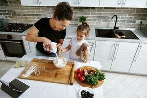 niña abrazos su felpa juguete y mezclas harina, cuales su mamá vierte dentro un bol. madre y hija Cocinando juntos foto